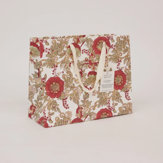 A square paper shopping bag with a vintage floral pattern in red, green, and gold on a white background, featuring cream-colored paper handles and a hanging tag.