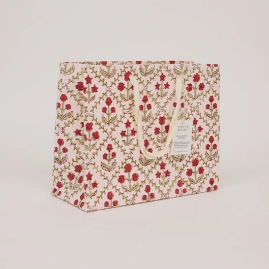 A square gift bag with a cream background and an all-over botanical pattern in red and olive green, featuring cream rope handles and a small white label on the front.