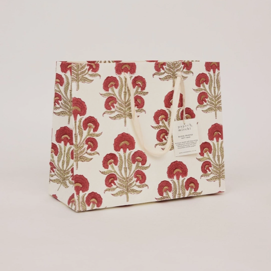 A paper shopping or gift bag with a cream background and a repeating red and gold botanical block print pattern, featuring cream paper handles and a small white brand label.