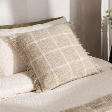 A square beige throw pillow with a white windowpane plaid pattern and fringed edges, styled on a cream-colored bed.