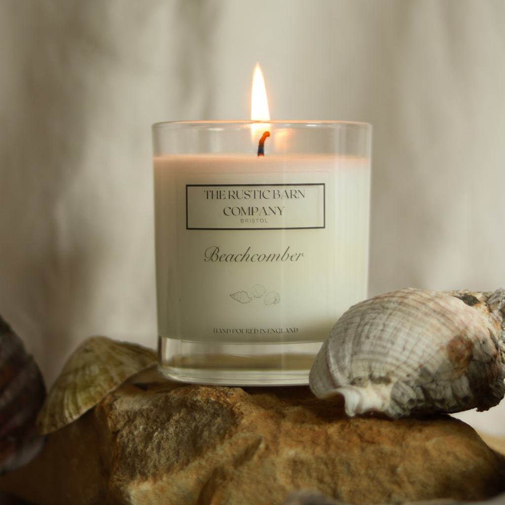 Beachcomber 220g candle by The Rustic Barn Company with a lit flame, displayed on coastal rocks with a seashell against a linen background