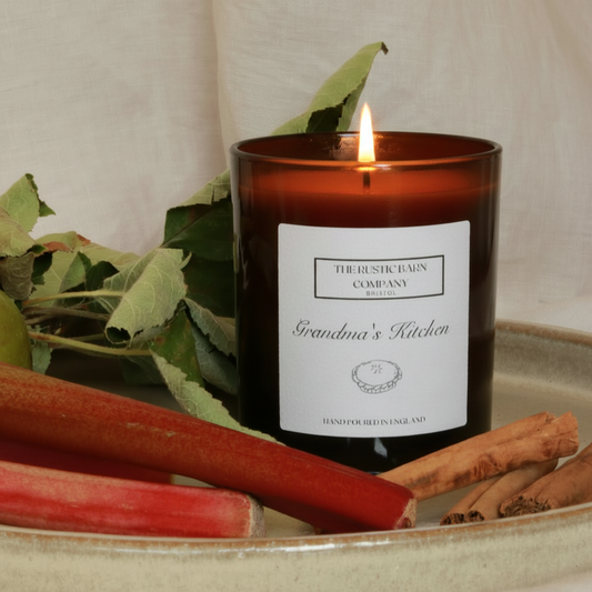 Grandma's Kitchen scented candle by The Rustic Barn Company, amber glass jar with lit flames, styled on a ceramic tray with fresh rhubarb stalks, cinnamon sticks and green leaves