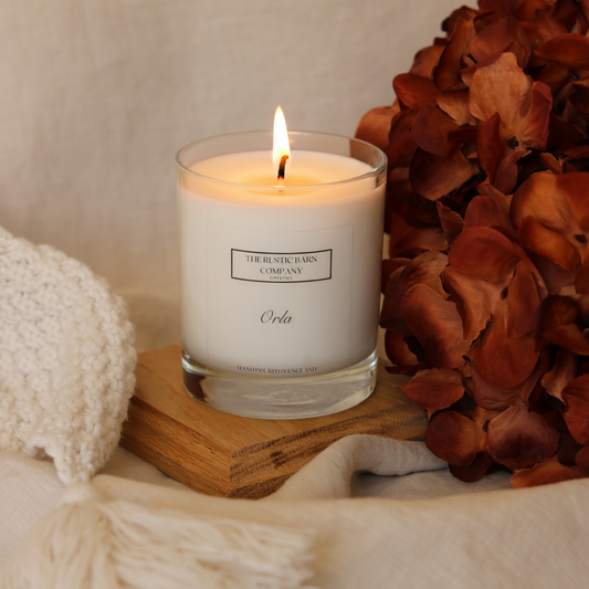 Orla scented candle by The Rustic Barn Company, clear glass jar on a wooden board styled with burnt orange dried hydrangeas and cream knit