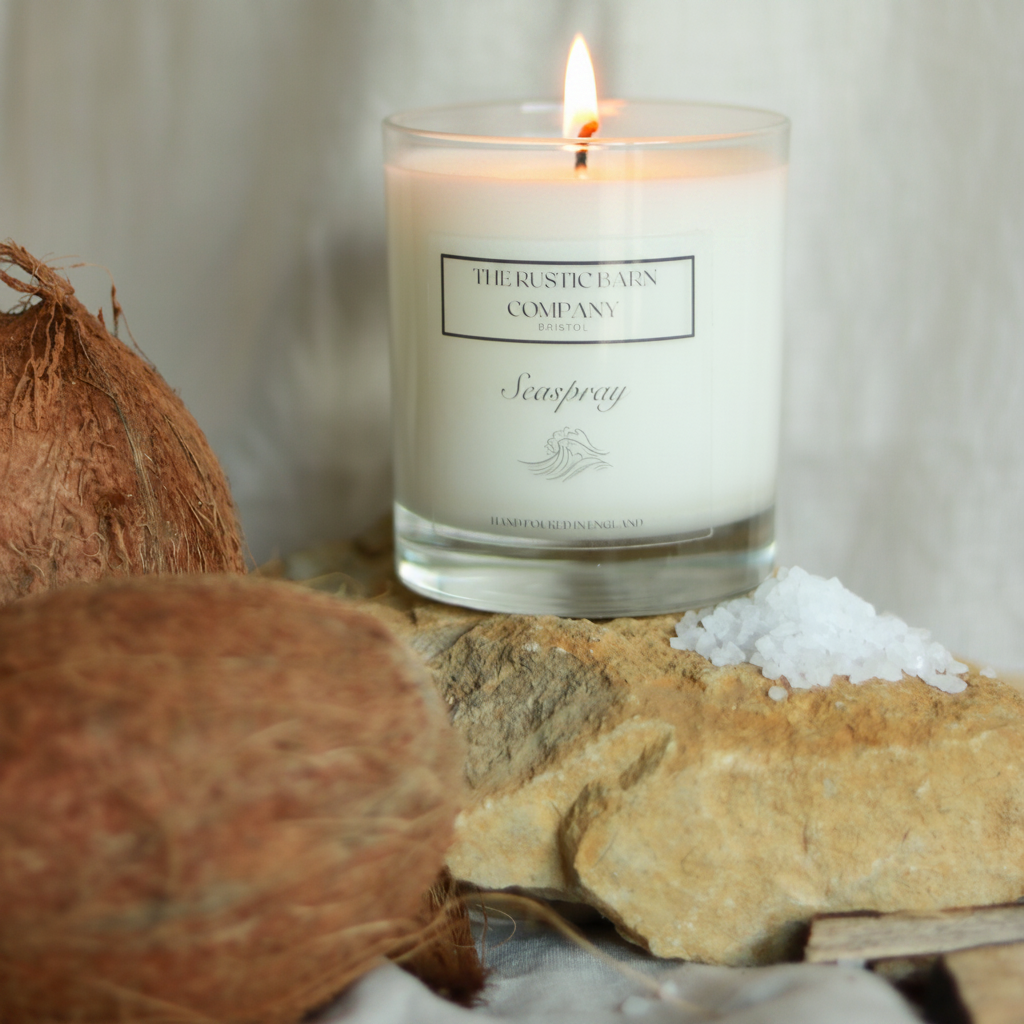 Seaspray 220g candle by The Rustic Barn Company with a lit flame, styled on coastal rocks with coconut halves, sea salt crystals and driftwood on a linen background
