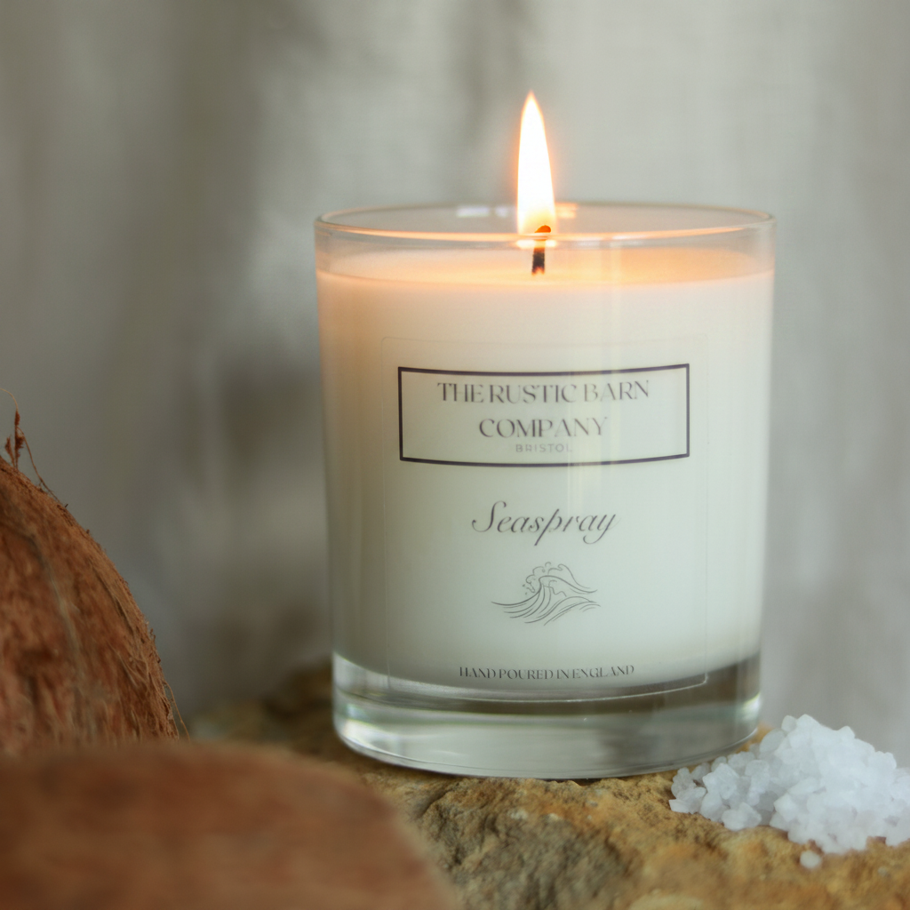 Seaspray 220g candle by The Rustic Barn Company with a lit flame, displayed on coastal rocks with a coconut half and sea salt crystals on a linen background
