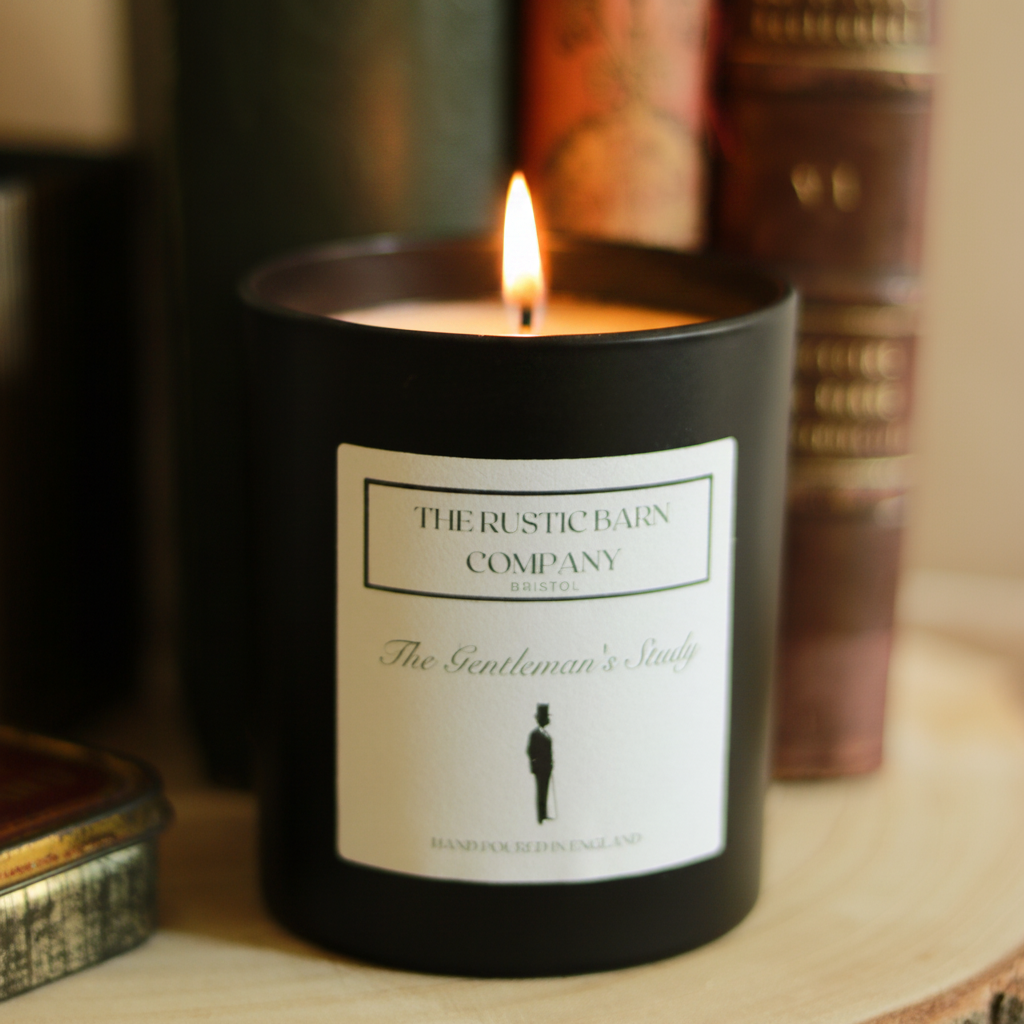 The Gentleman's Study 220g candle by The Rustic Barn Company with a lit flame, close-up shot on a wooden slice with leather-bound books softly blurred in the background