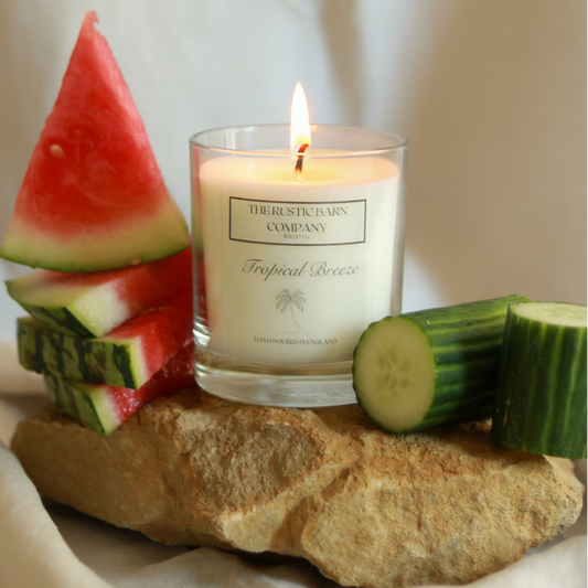 Tropical Breeze 220g candle by The Rustic Barn Company with a lit flame, displayed on rocks surrounded by fresh watermelon slices and cucumber on a linen background

