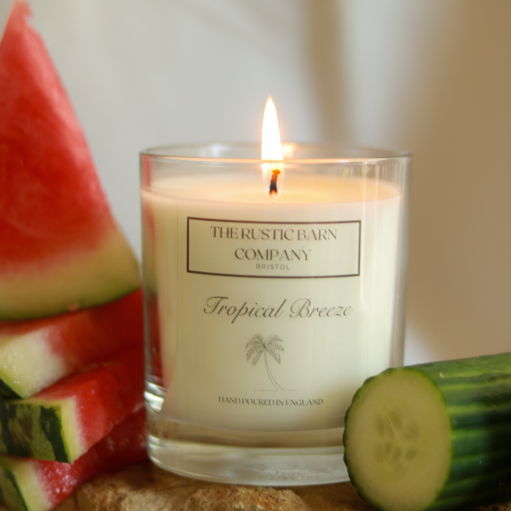 Tropical Breeze 220g candle by The Rustic Barn Company with a lit flame, close-up shot with watermelon wedges to the left and a sliced cucumber to the right on a warm neutral background